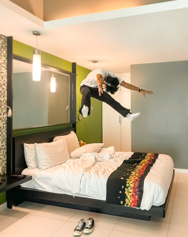 Person jumping playfully on a comfortable hotel bed with decorative pillows and modern lighting