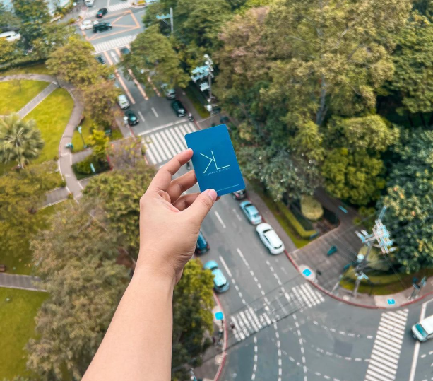 Hand holding a blue card with a white design over a city intersection with cars and trees.