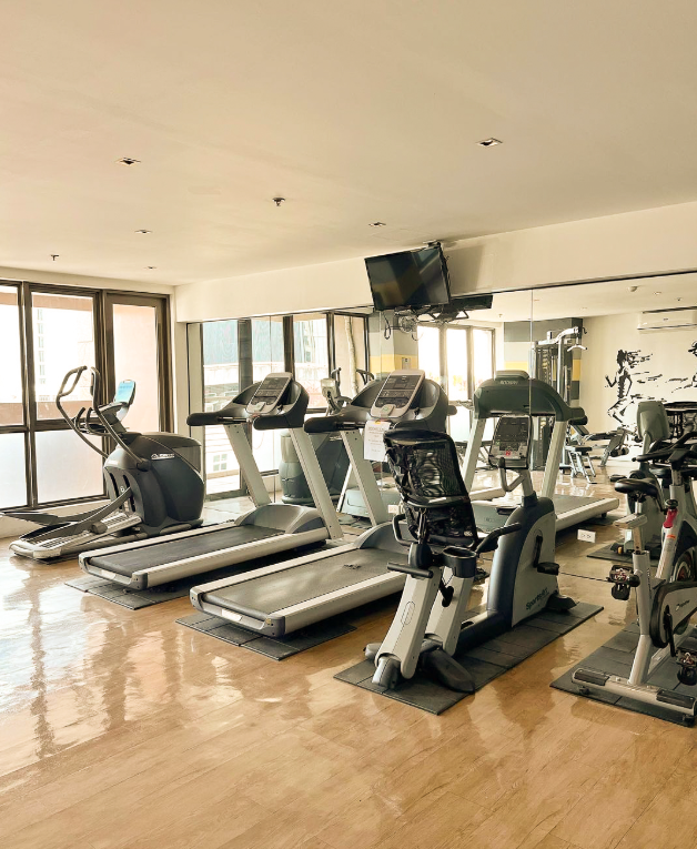 Gym interior with treadmills, elliptical, stationary bikes, and weights. Mirrored wall reflects equipment.