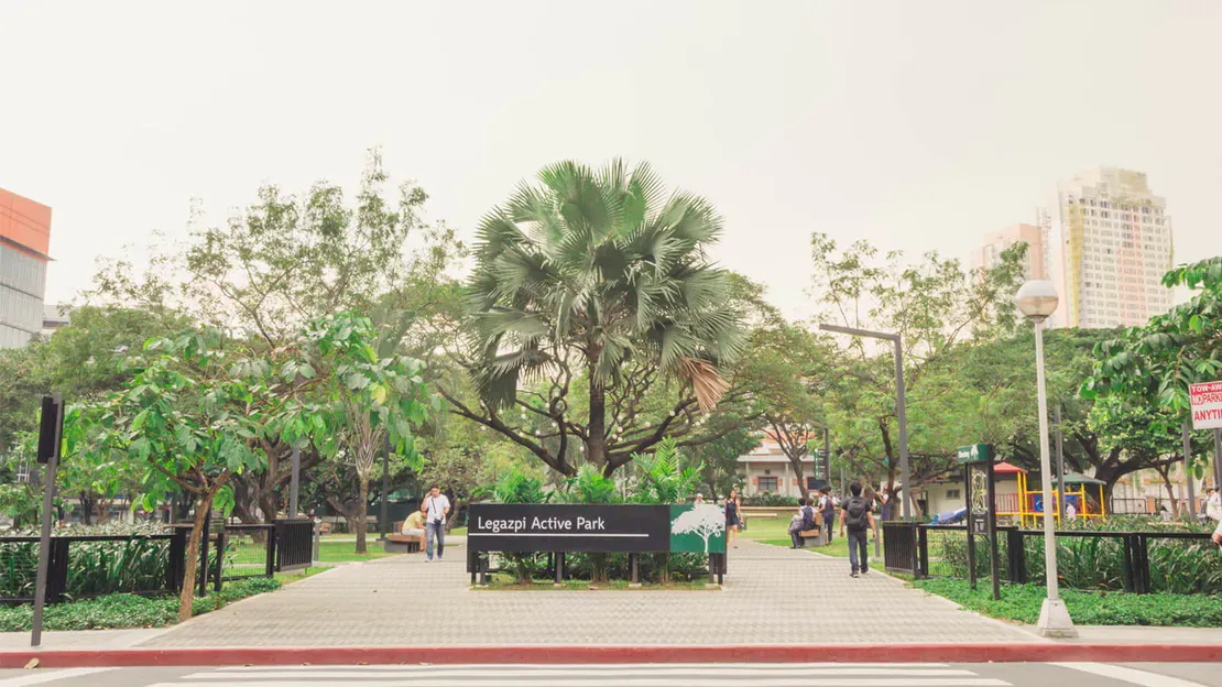 Tree-lined urban park with paved walkway, benches, and a distant city building in the background