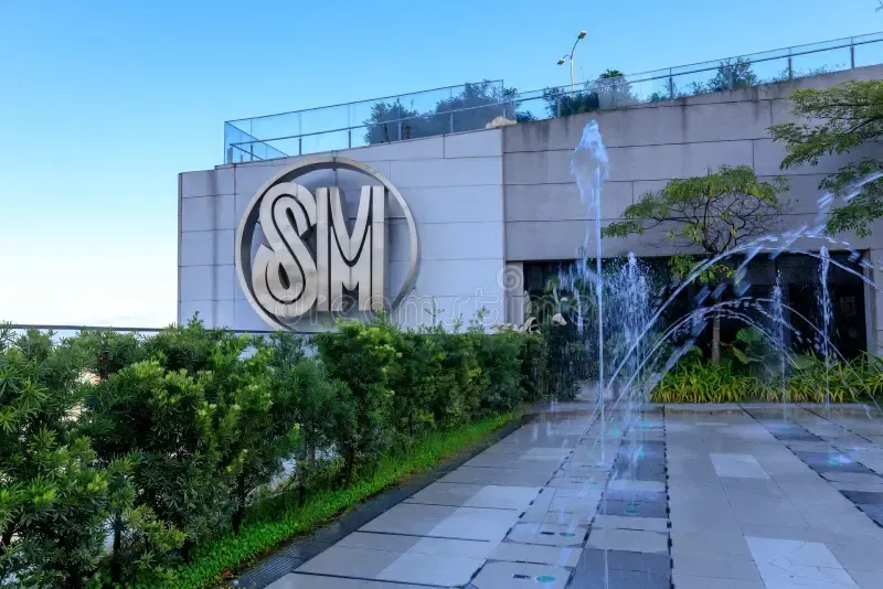 SM Mall exterior with large logo, fountain, and greenery against a blue sky.