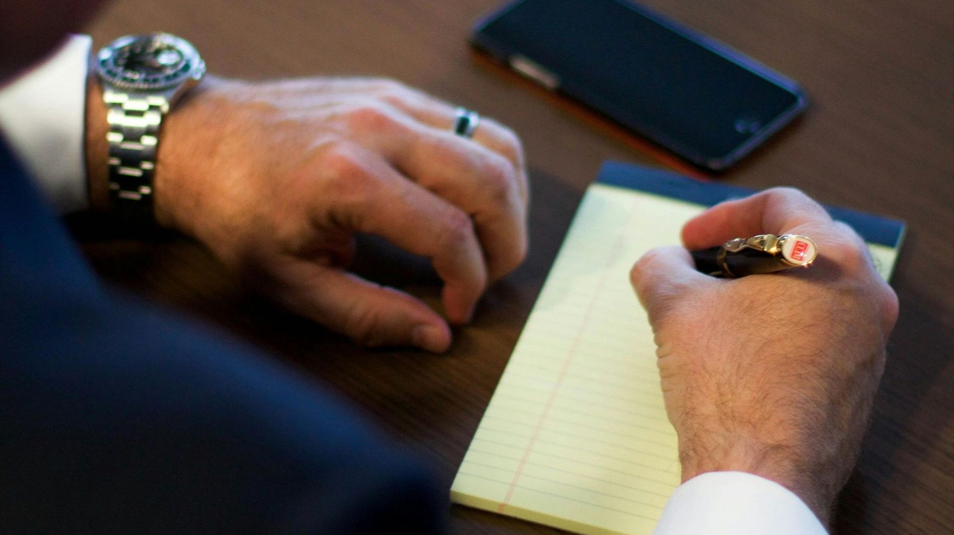 Hands writing on a notepad at a desk beside a smartphone.