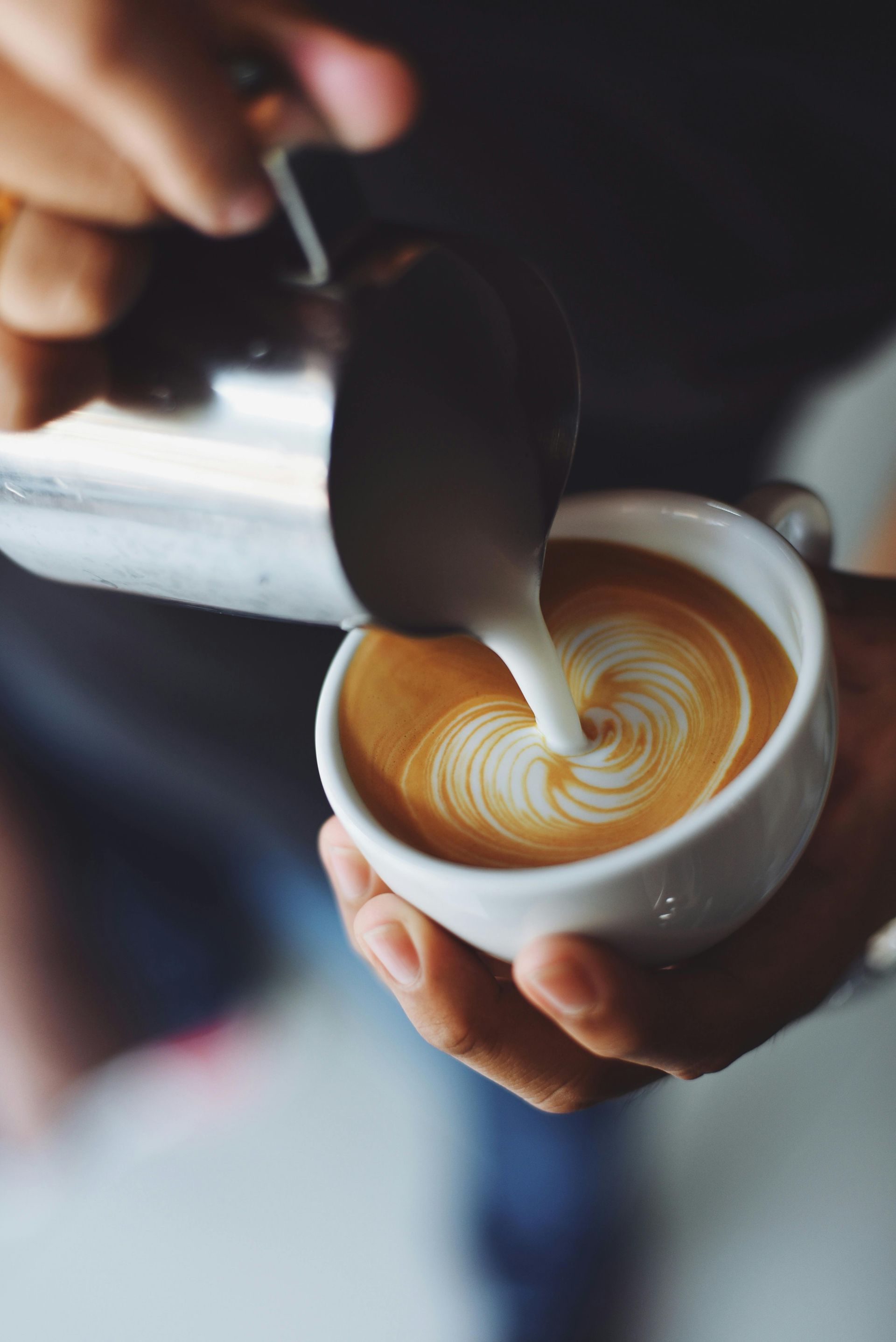 Barista pouring milk into coffee, creating a swirling latte art design in a white mug.