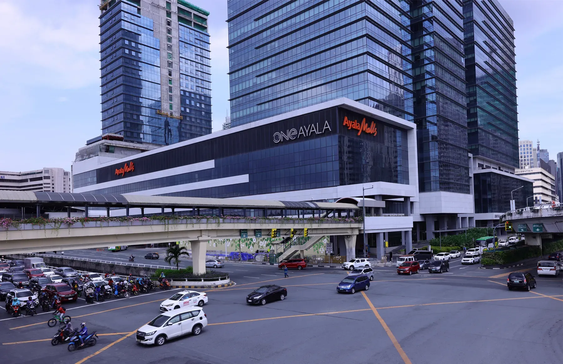 A busy intersection with cars and motorcycles in front of the modern, glass-paneled One Ayala commercial complex.
