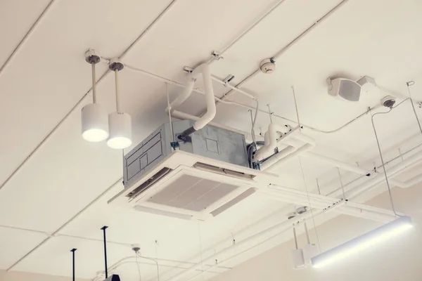 Un condizionatore d'aria a soffitto circondato da lampade a sospensione e cavi elettrici a vista in un interno in stile industriale.