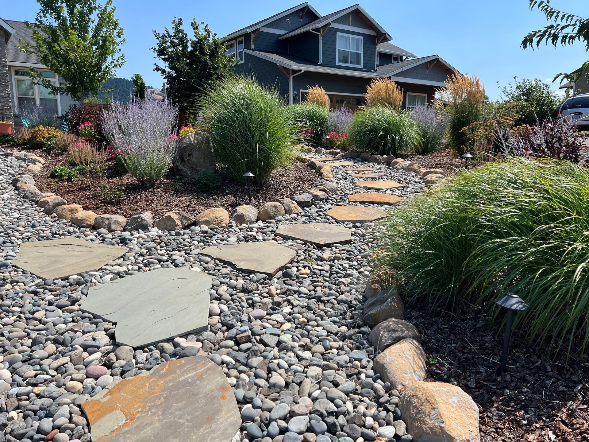 Stone path through a landscaped front yard leading to a blue house under a clear sky