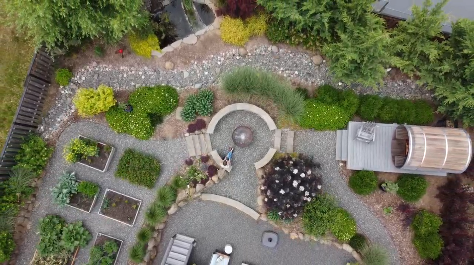 Aerial view of a landscaped garden with stone paths, circular patio, planters, and a greenhouse shed