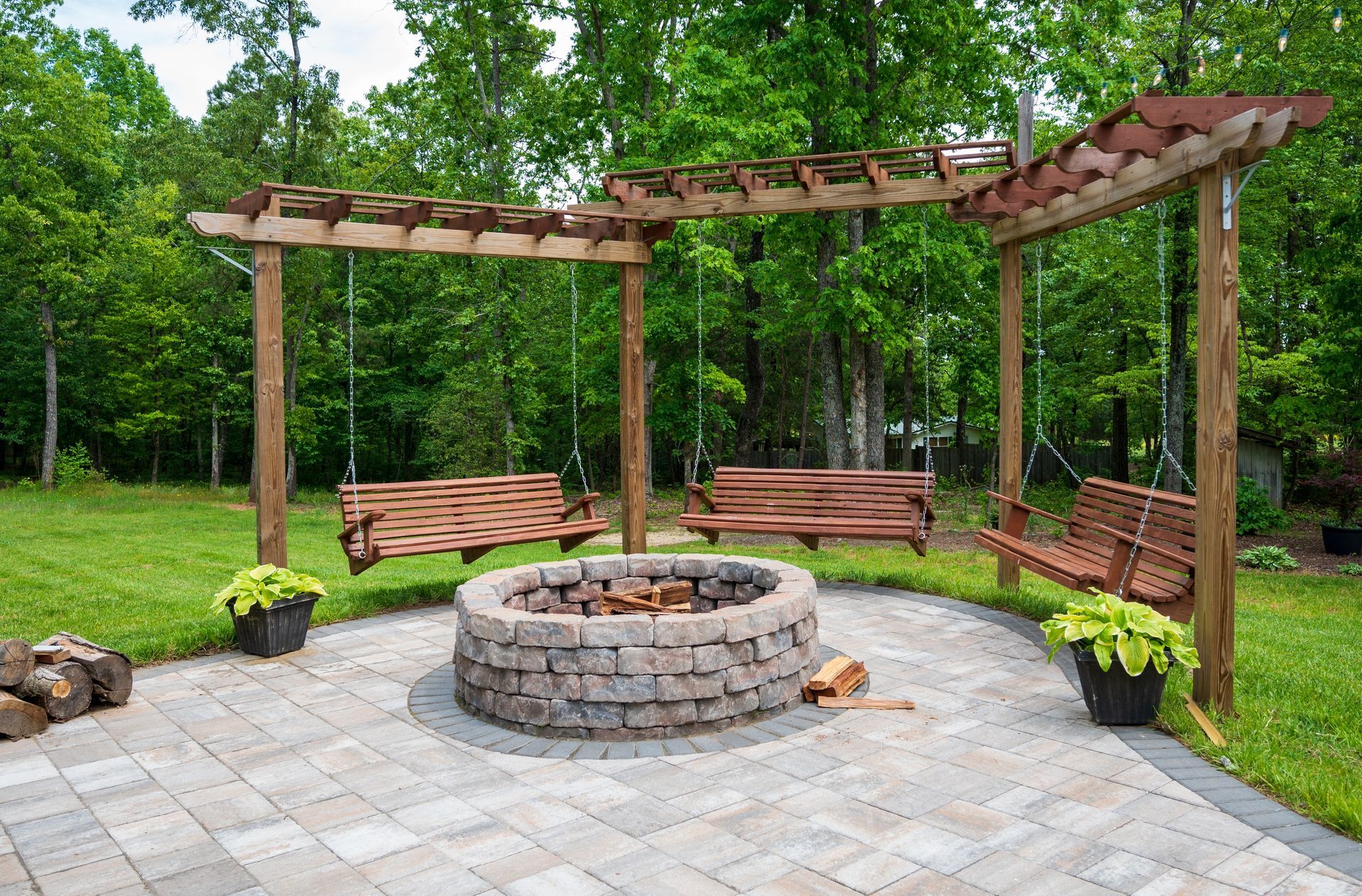 Backyard fire pit with curved wooden benches under a pergola on a stone patio surrounded by trees