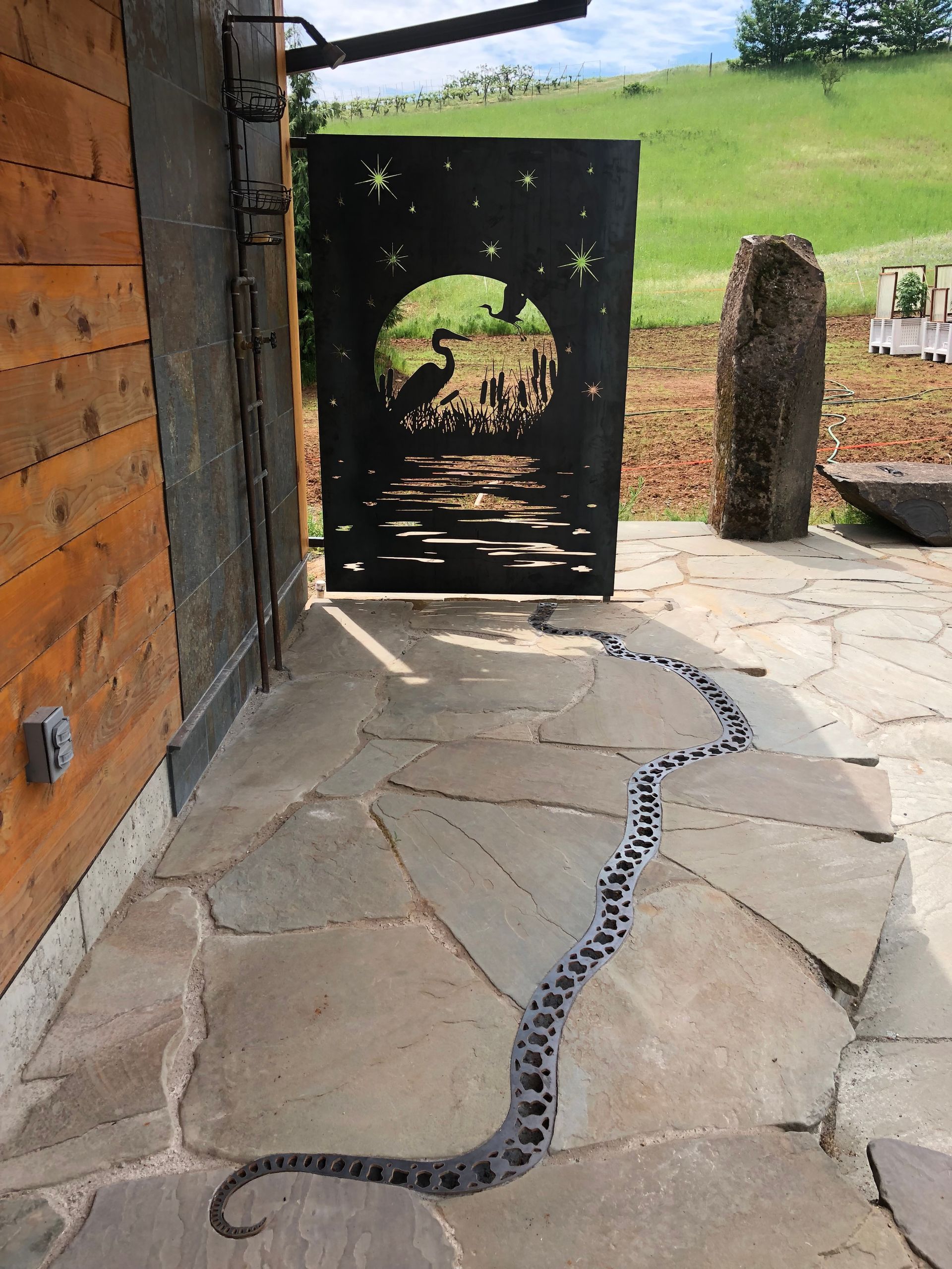 Stone patio with decorative snake leads to a black metal gate with a landscape silhouette; a building and grassy hill are in the background.