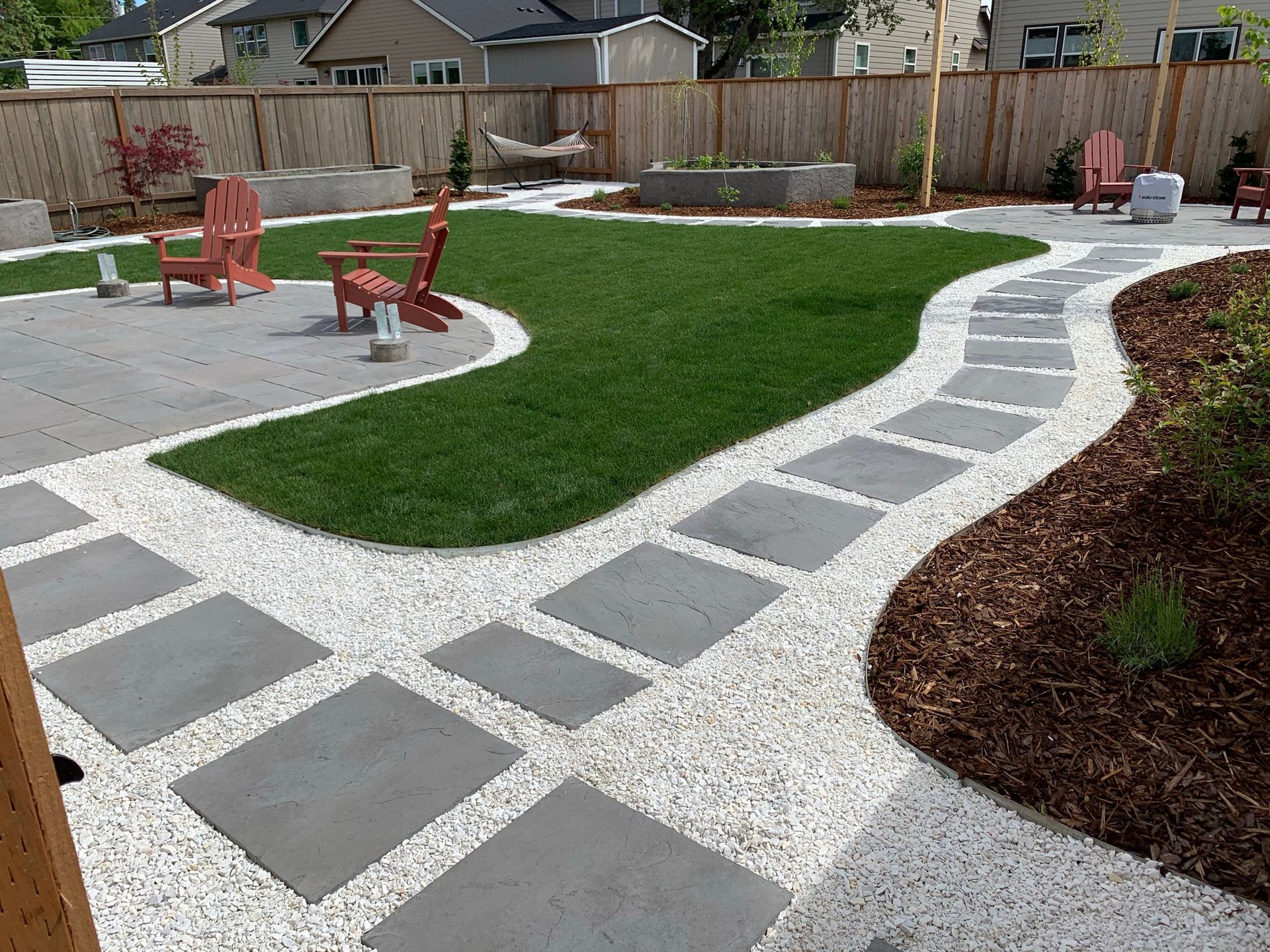 Backyard patio with stepping stones, green lawn, gravel, wooden fence, and chairs.