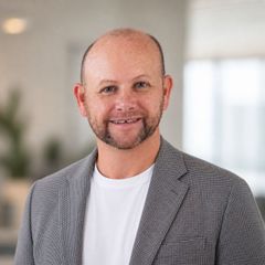 Jason Pascoe in a red plaid blazer over a white shirt, standing against a white brick wall with a slight smile.
