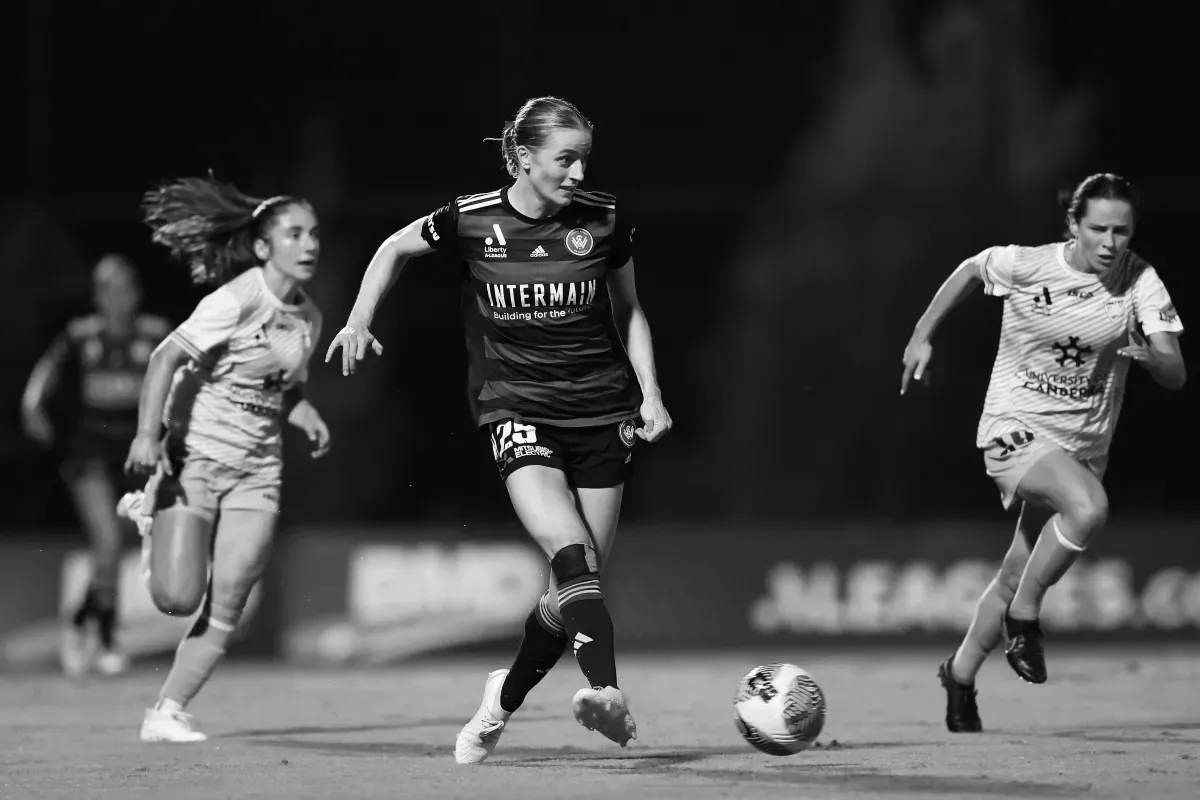 Female soccer players in action on a field. One dribbles the ball, two others chase in the background.