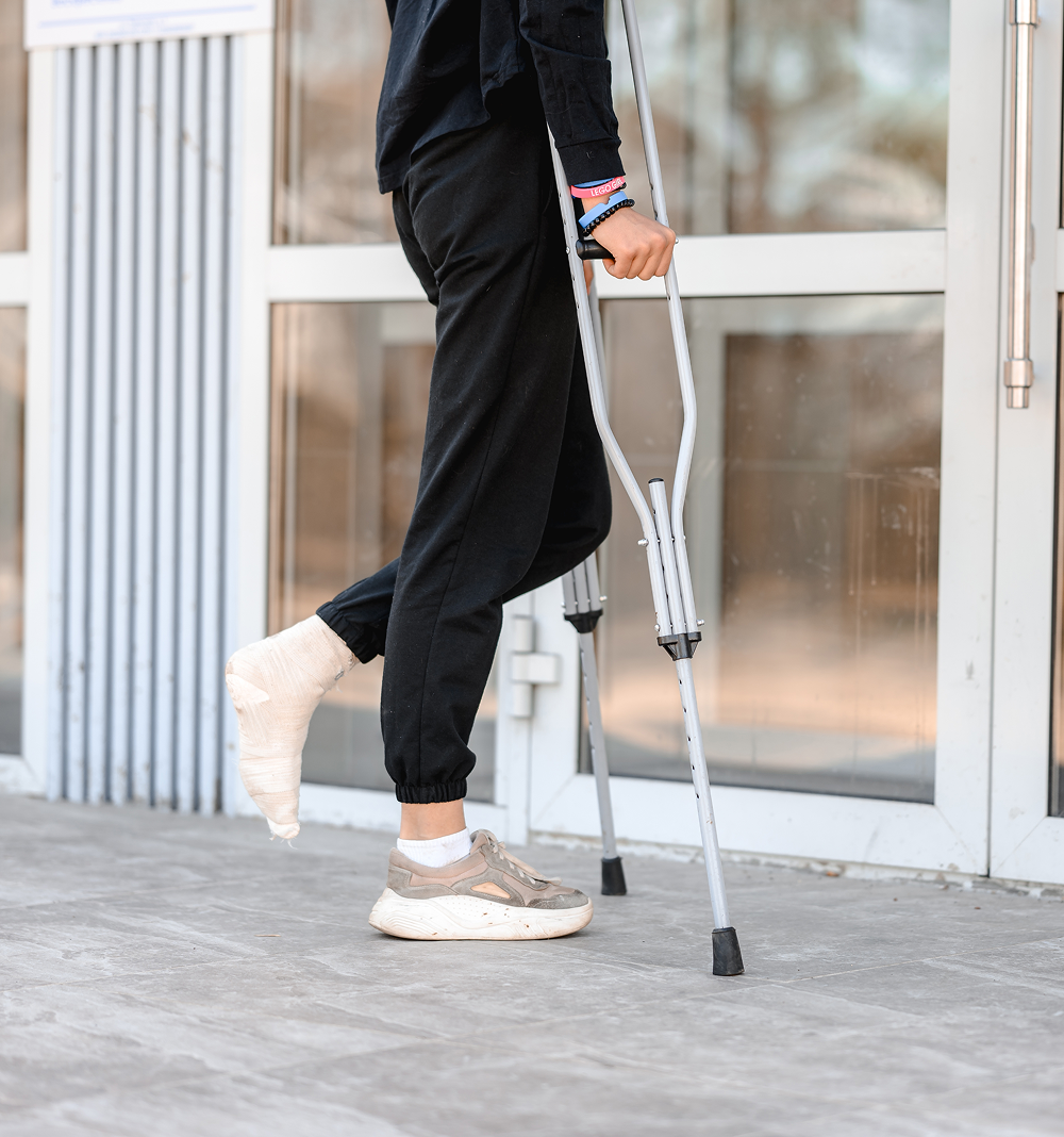 Person wearing a cast on their left foot using crutches to walk near glass doors.