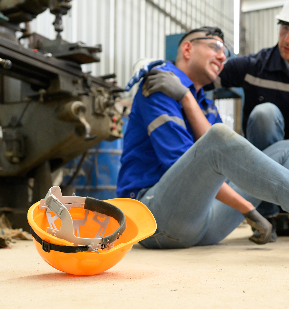 Worker injured on factory floor, assisted by another worker, safety helmet in foreground.