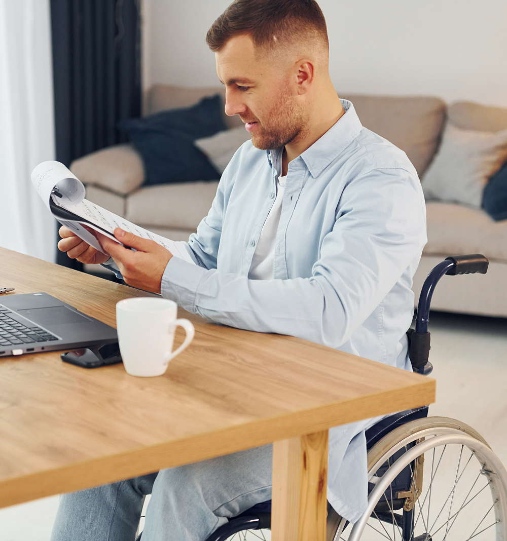 A man in a wheelchair, working at a wooden desk with a laptop, mug, and documents.
