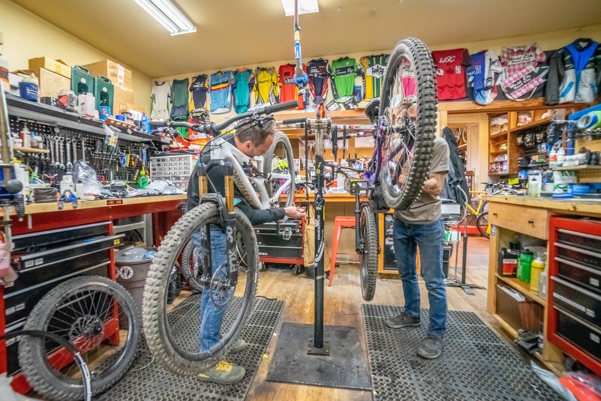 Bike repair shop with a mechanic fixing a bicycle on a stand amid tools and hanging gear.