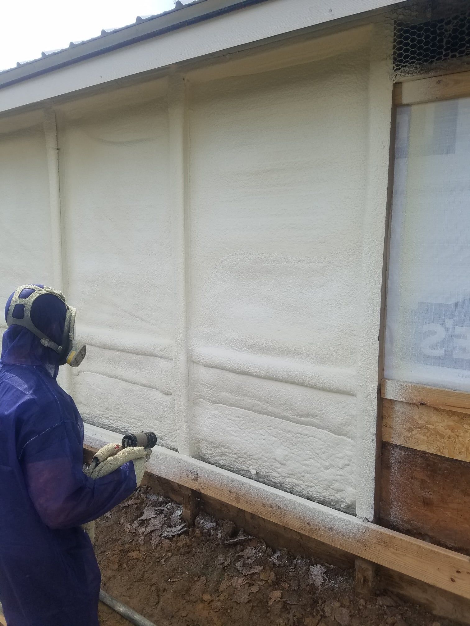 Person in protective gear spraying foam insulation onto a building's exterior wall.
