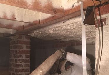Worker in protective suit spraying insulation into a crawl space, brick supports and wooden beams visible.