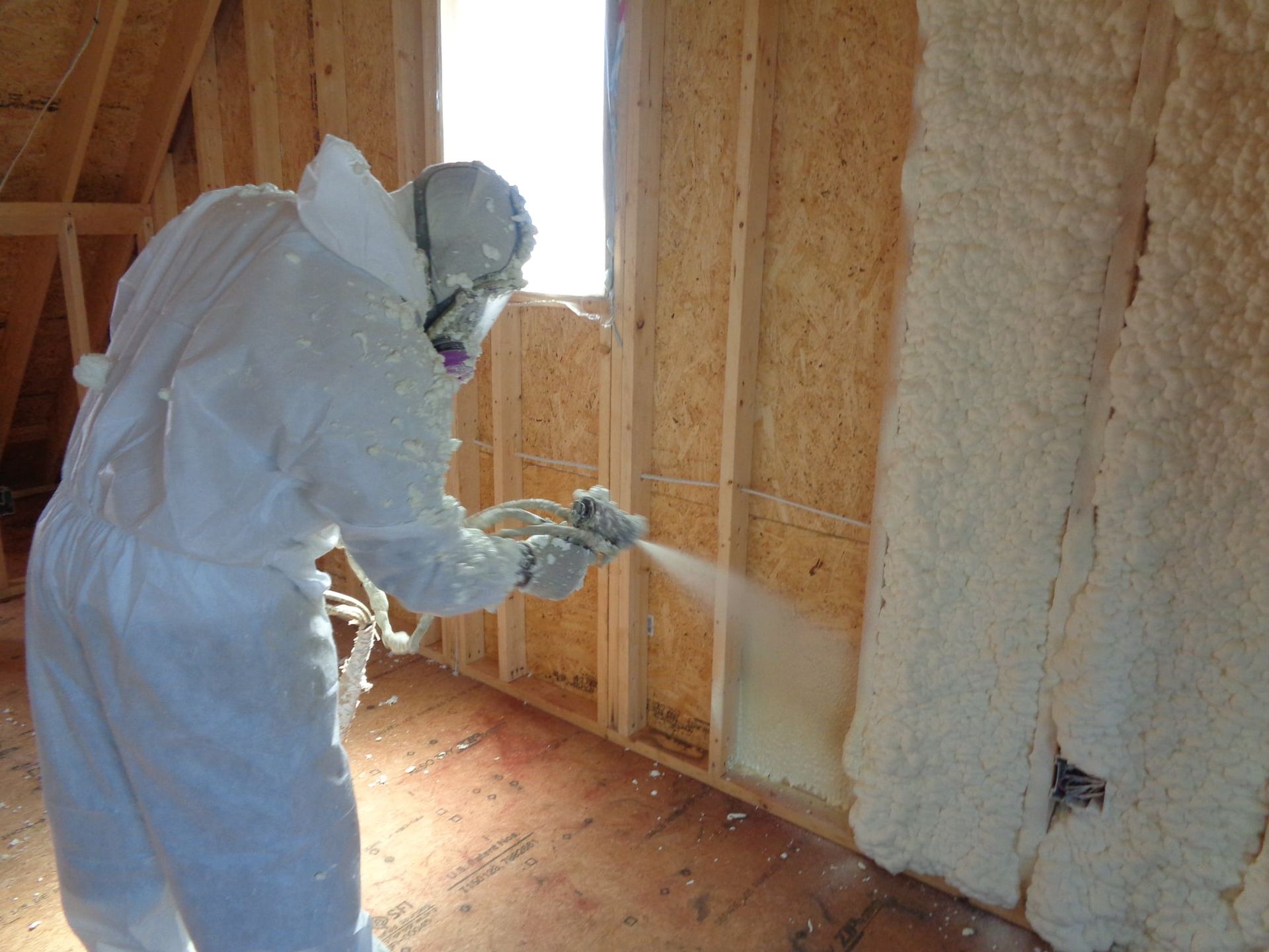 Person in protective suit spraying insulation foam inside a wooden-framed building.