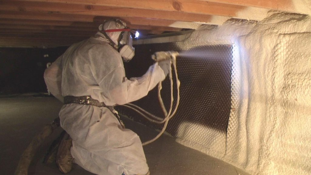 Person in protective suit spraying foam insulation in a crawl space.
