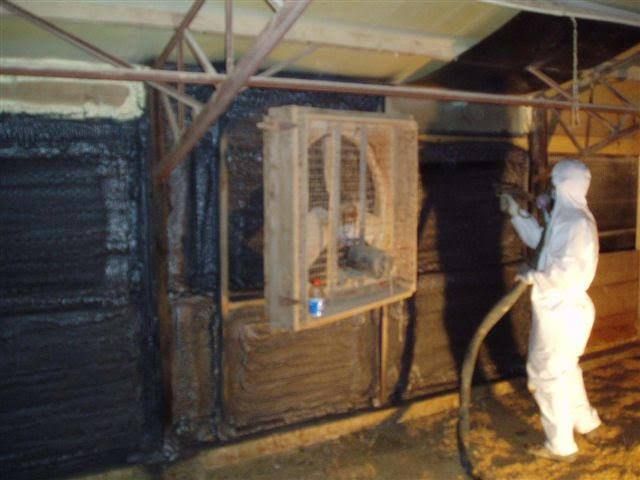 Person in protective suit spraying insulation on interior wall; ventilation unit visible.