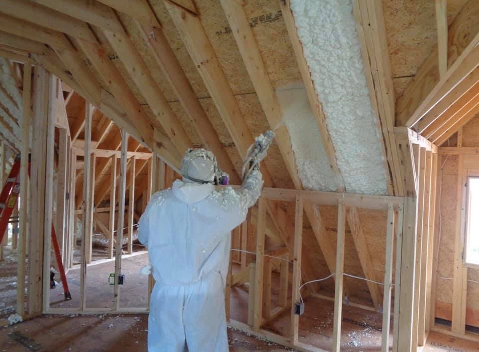 Person in protective suit spraying foam insulation in a wooden house frame.
