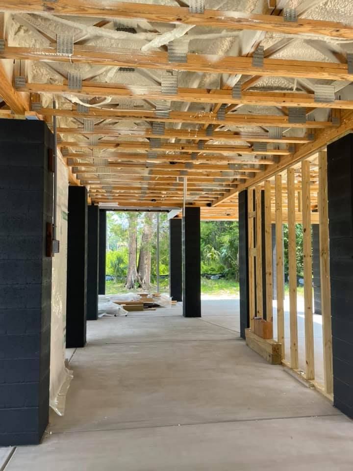 Under-construction porch with exposed wooden beams, concrete floor, black columns, and wooded background.