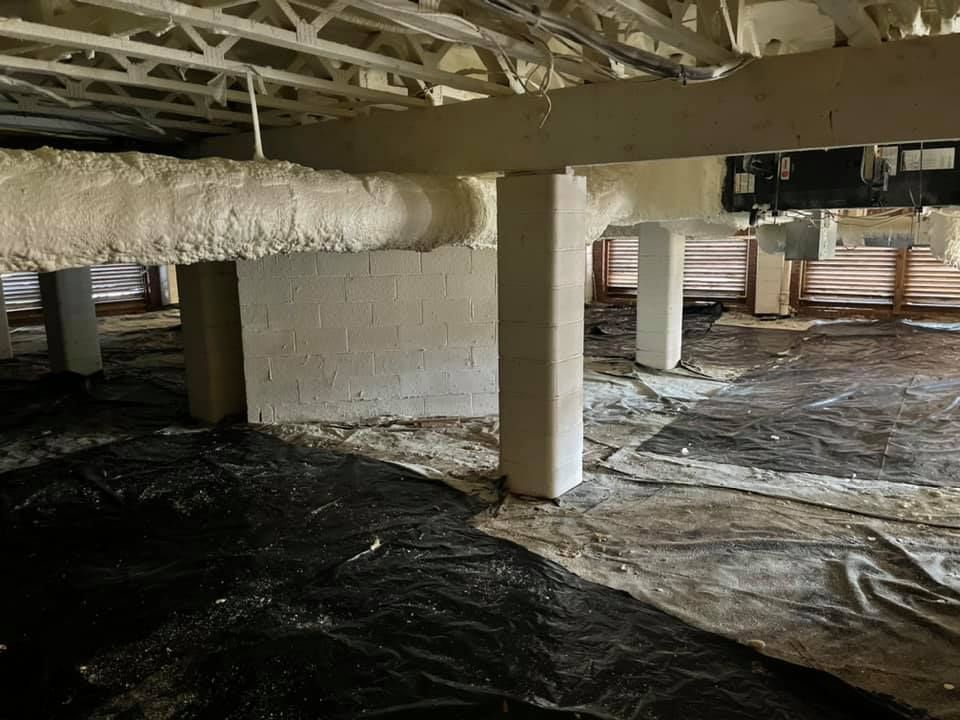 Crawlspace with insulation sprayed on ductwork and supports, plastic sheeting on floor. White and black.