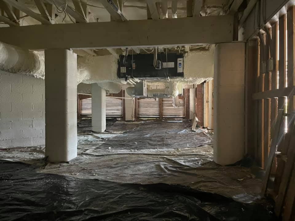Interior view of a building under renovation, with exposed beams, spray foam insulation, and a central HVAC unit.