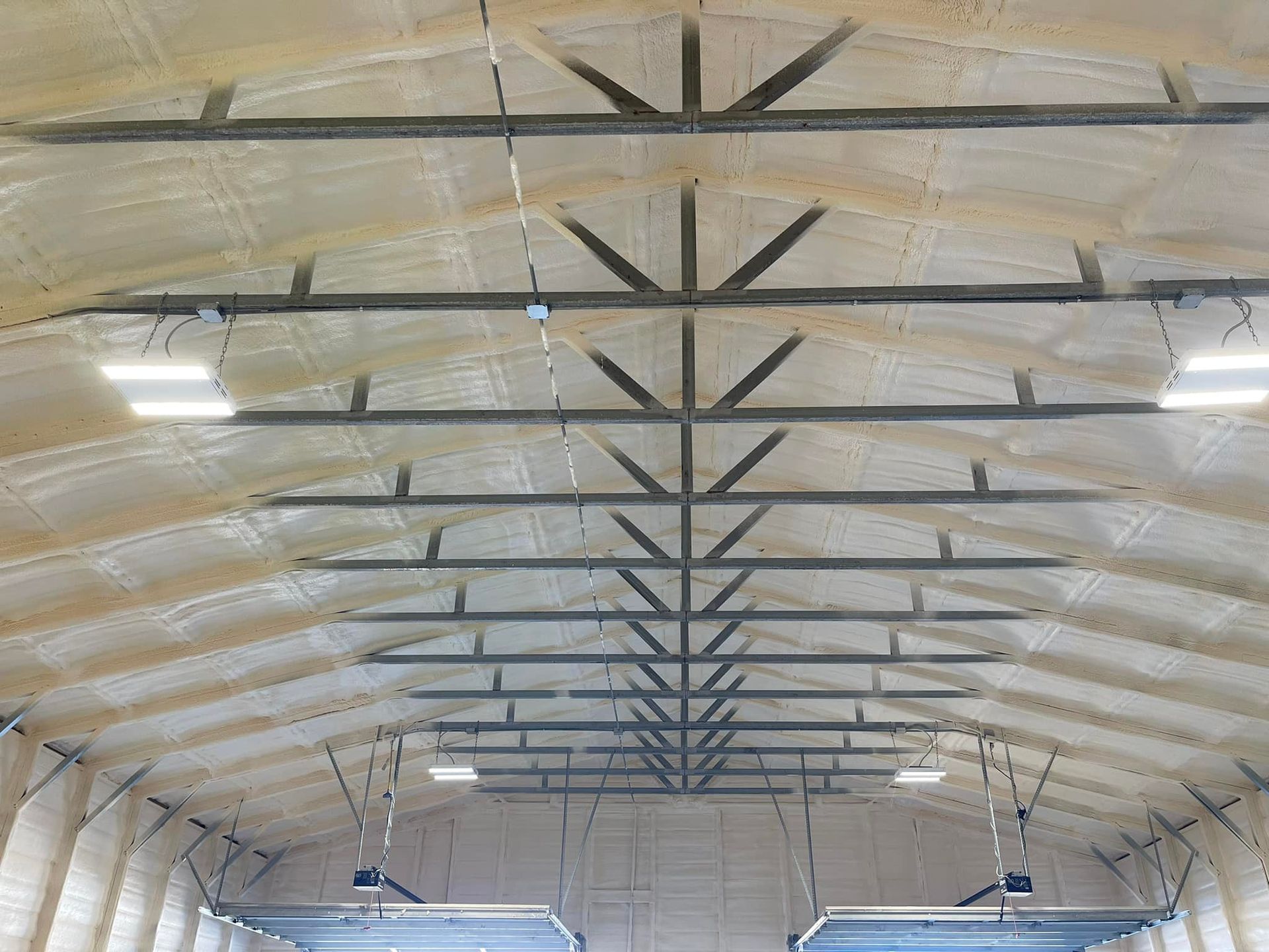 Interior of a building with a truss roof and foam insulation. Overhead lights are visible.