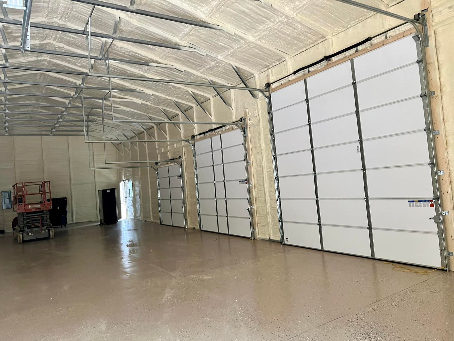 Interior view of a warehouse with large white garage doors, spray foam insulation, and metal framework.