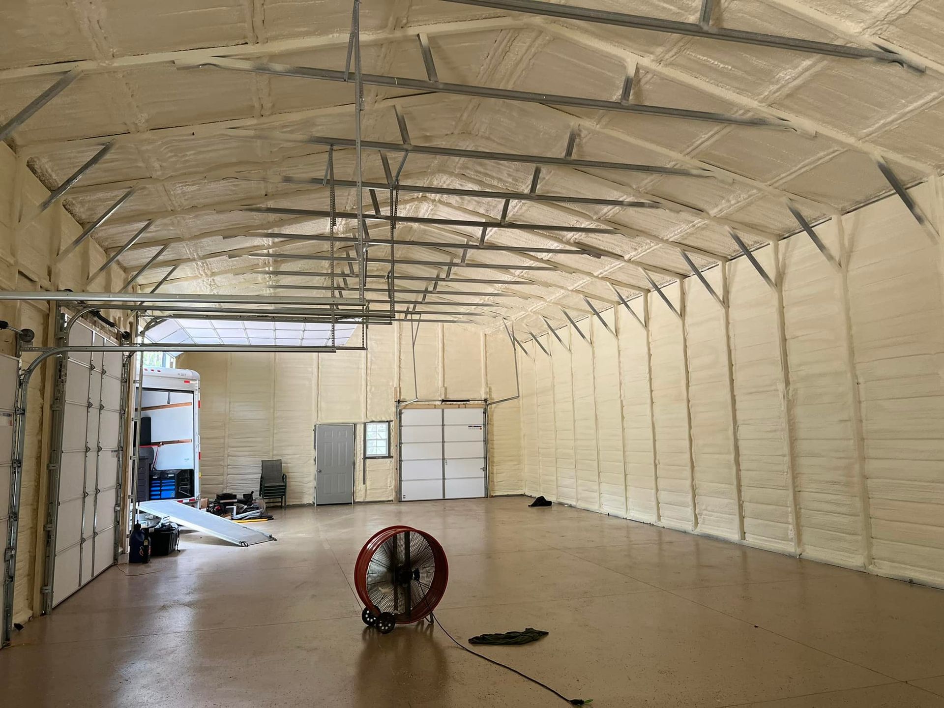 Interior of a building with light tan spray foam insulation on the walls and ceiling, a large fan, and a shiny floor.