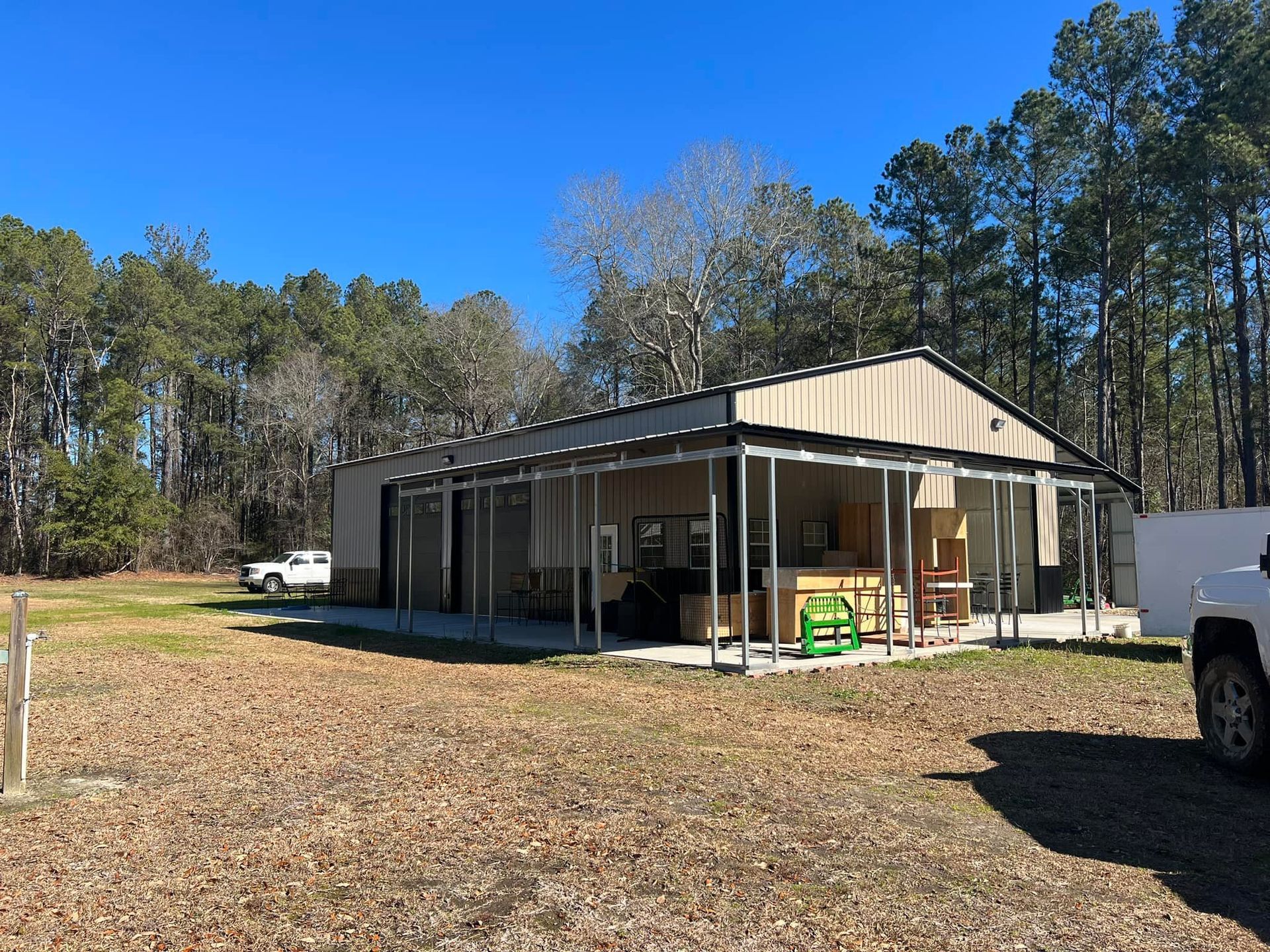 Metal shed with a covered porch on a grassy lot, surrounded by trees; two trucks parked nearby.