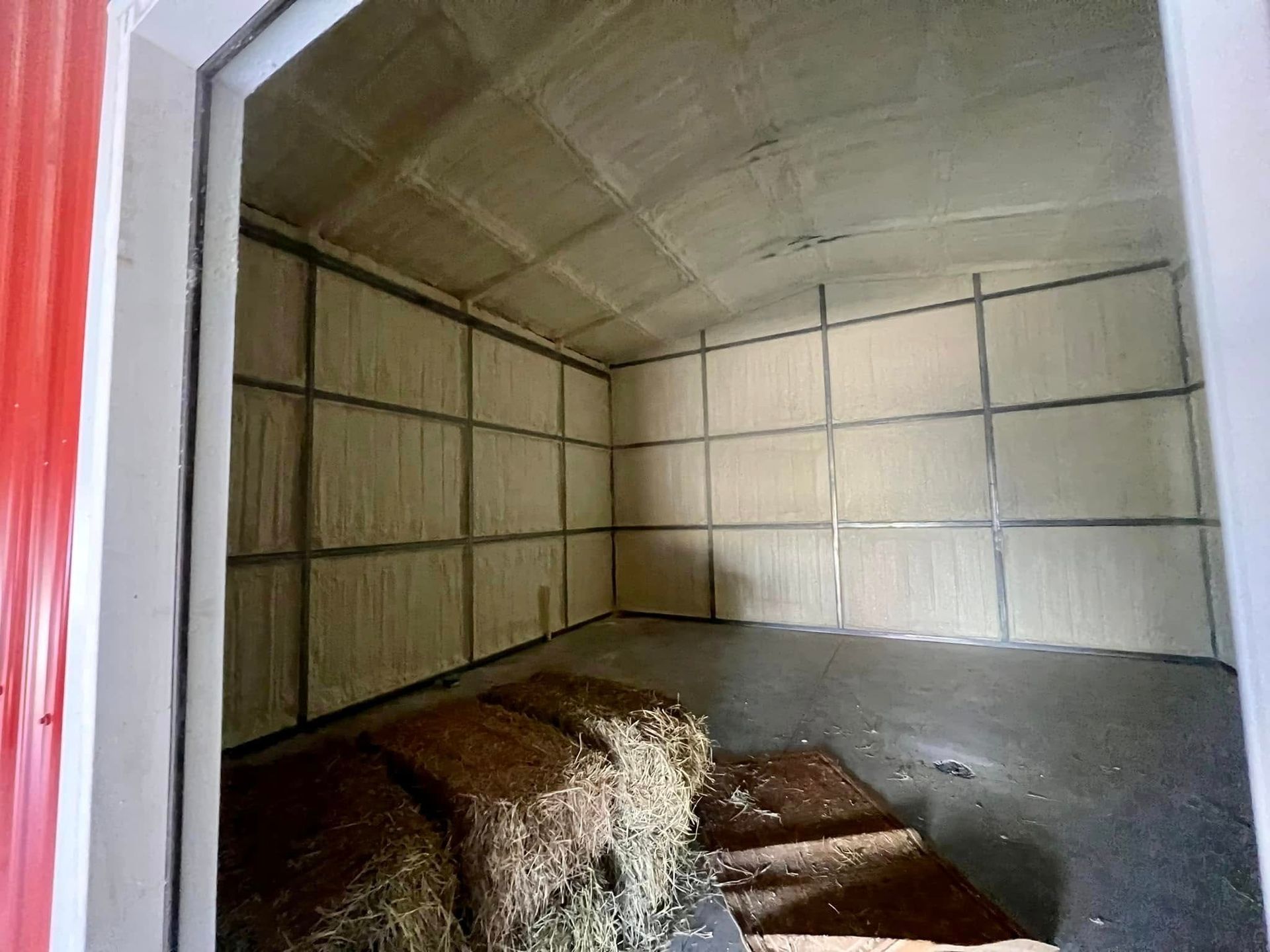 Interior of a shed, insulated with light-colored material and metal grid, hay bales in foreground.