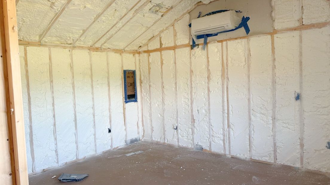 Interior of a room with spray foam insulation between wooden studs, and an air conditioning unit on the wall.