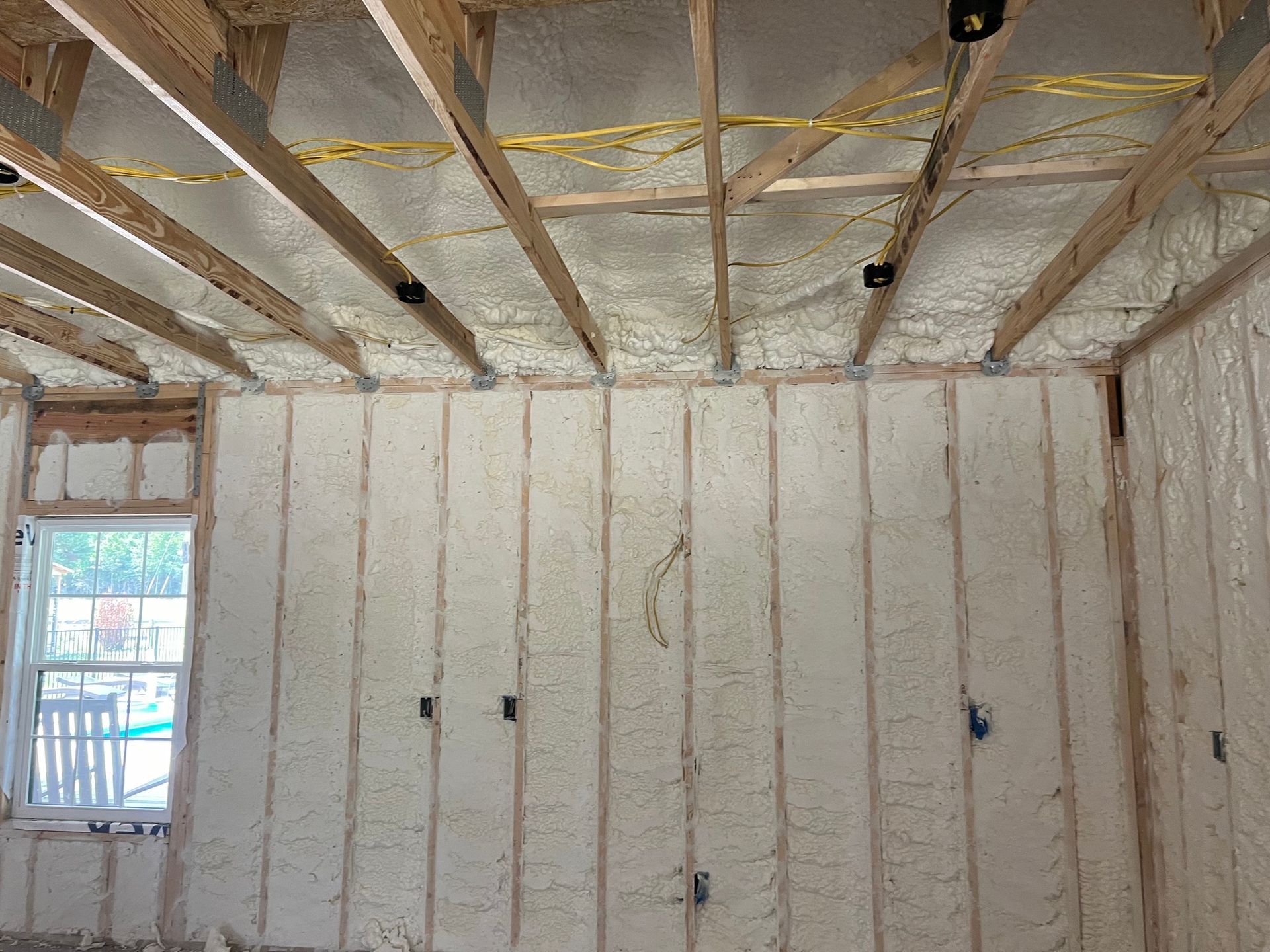 Interior view of a room under construction, with exposed wooden beams and spray foam insulation on the walls and ceiling.