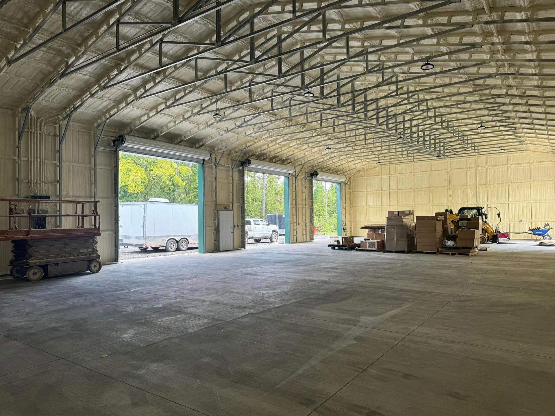 Inside of a large warehouse with arched roof, three open garage doors. Heavy equipment and boxes are visible.