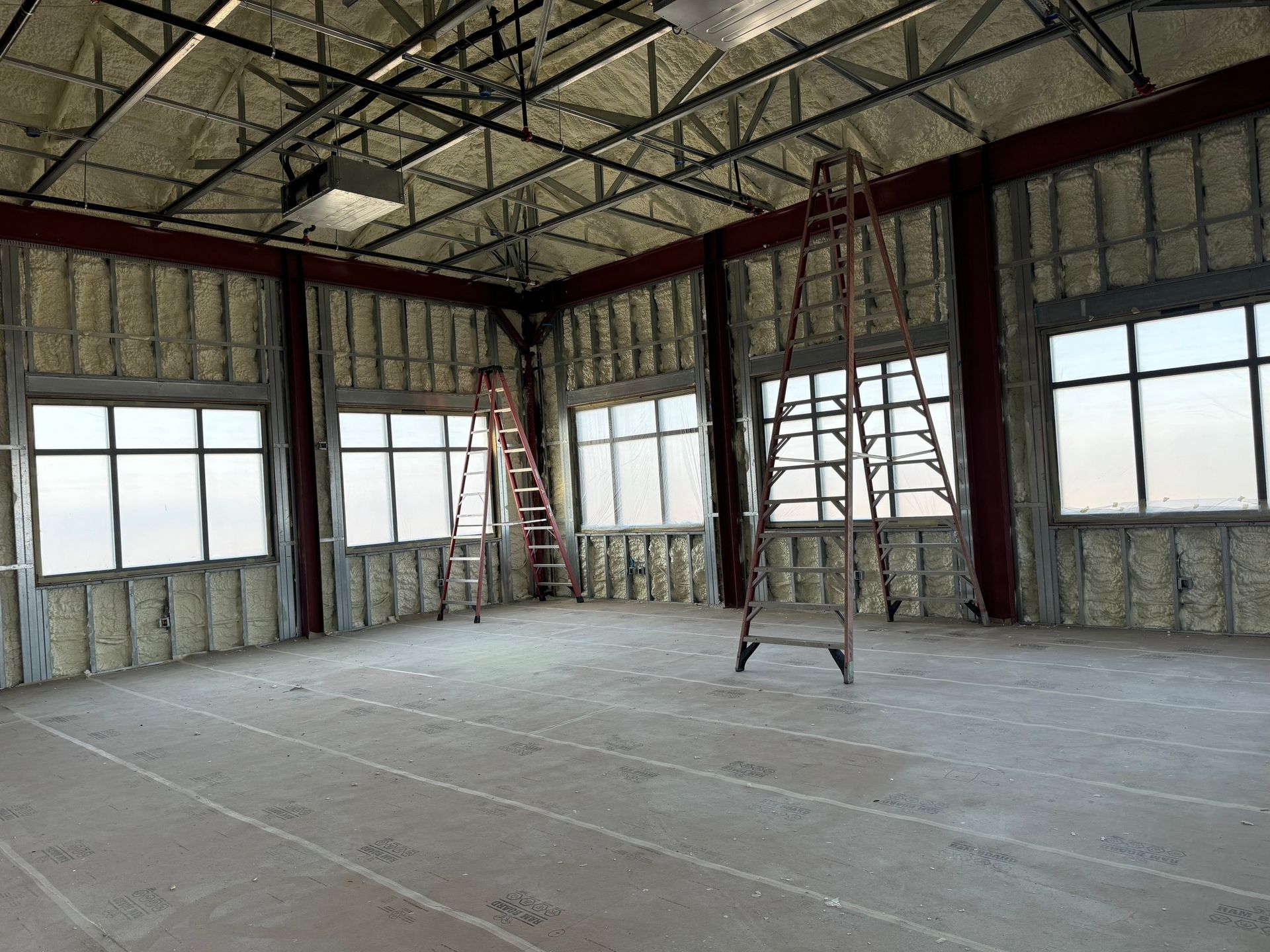Interior of a building under construction, windows, ladders, exposed metal framework, and concrete floor.
