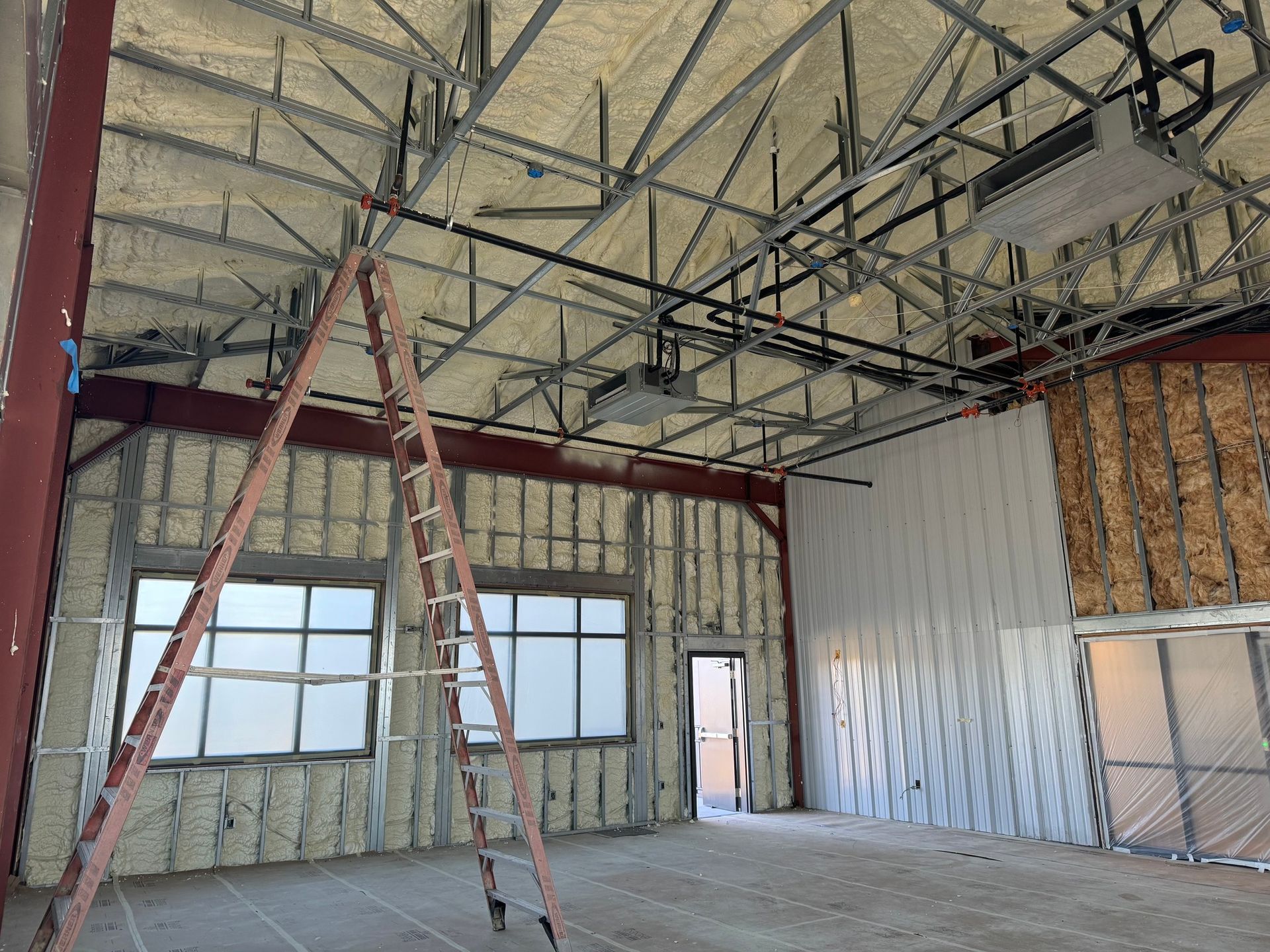 Interior view of a building under construction, featuring exposed steel beams, spray foam insulation, and a ladder.