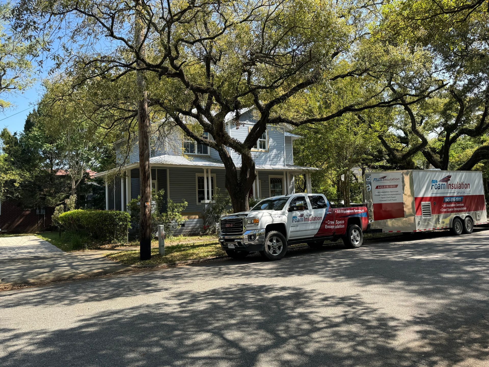 A truck towing a moving trailer parked in front of a light blue two-story house with a large tree.