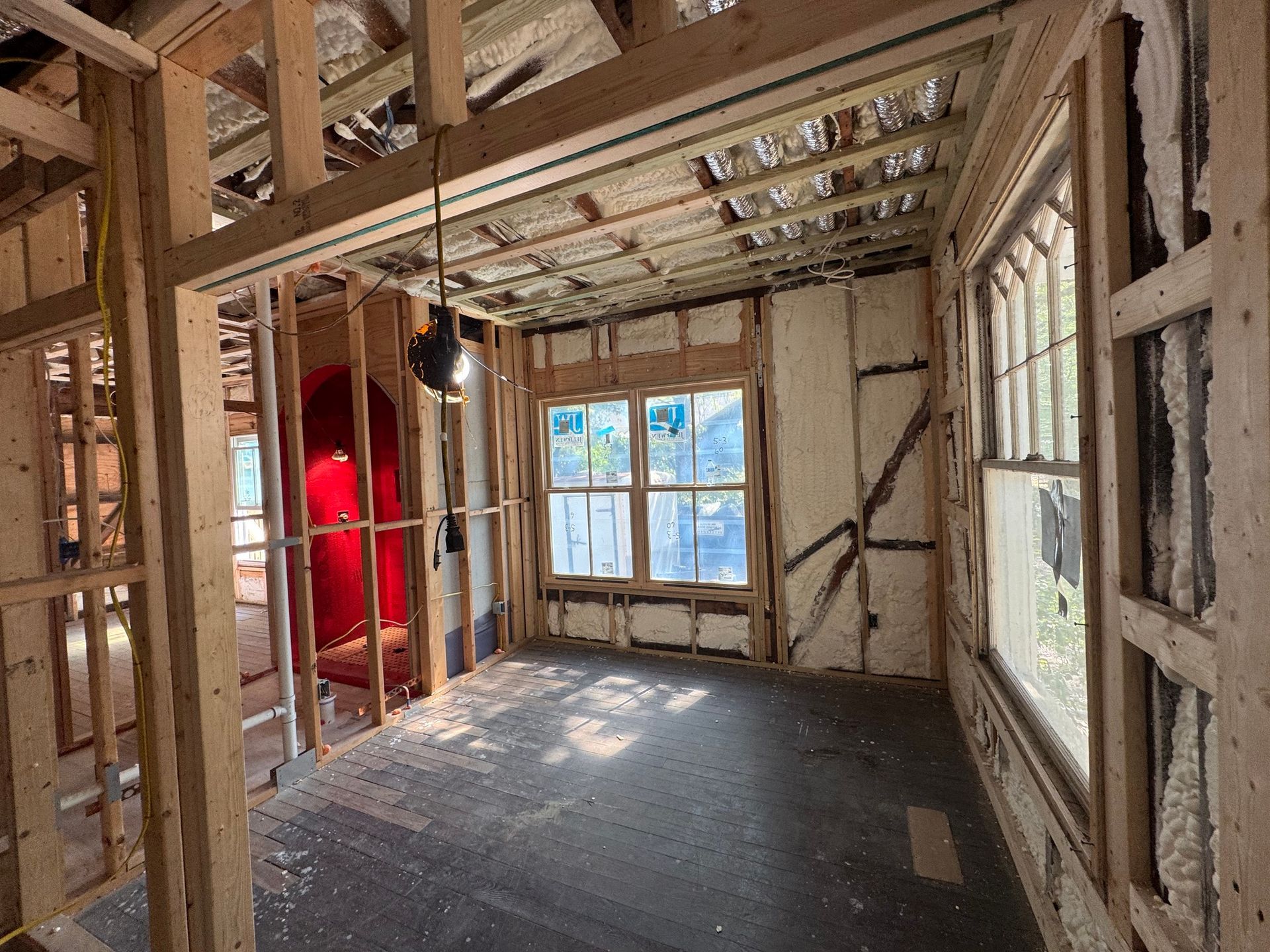 Interior room under construction, exposed wooden framing, insulation visible, windows, dark floor.