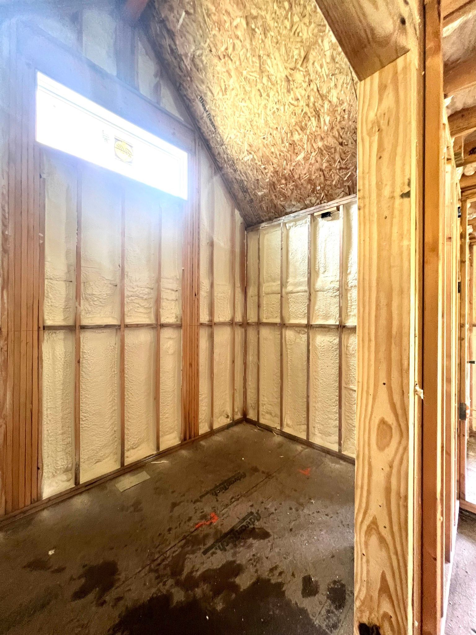 Interior of a building under construction, walls and ceiling insulated with foam, wood framing visible, natural light.