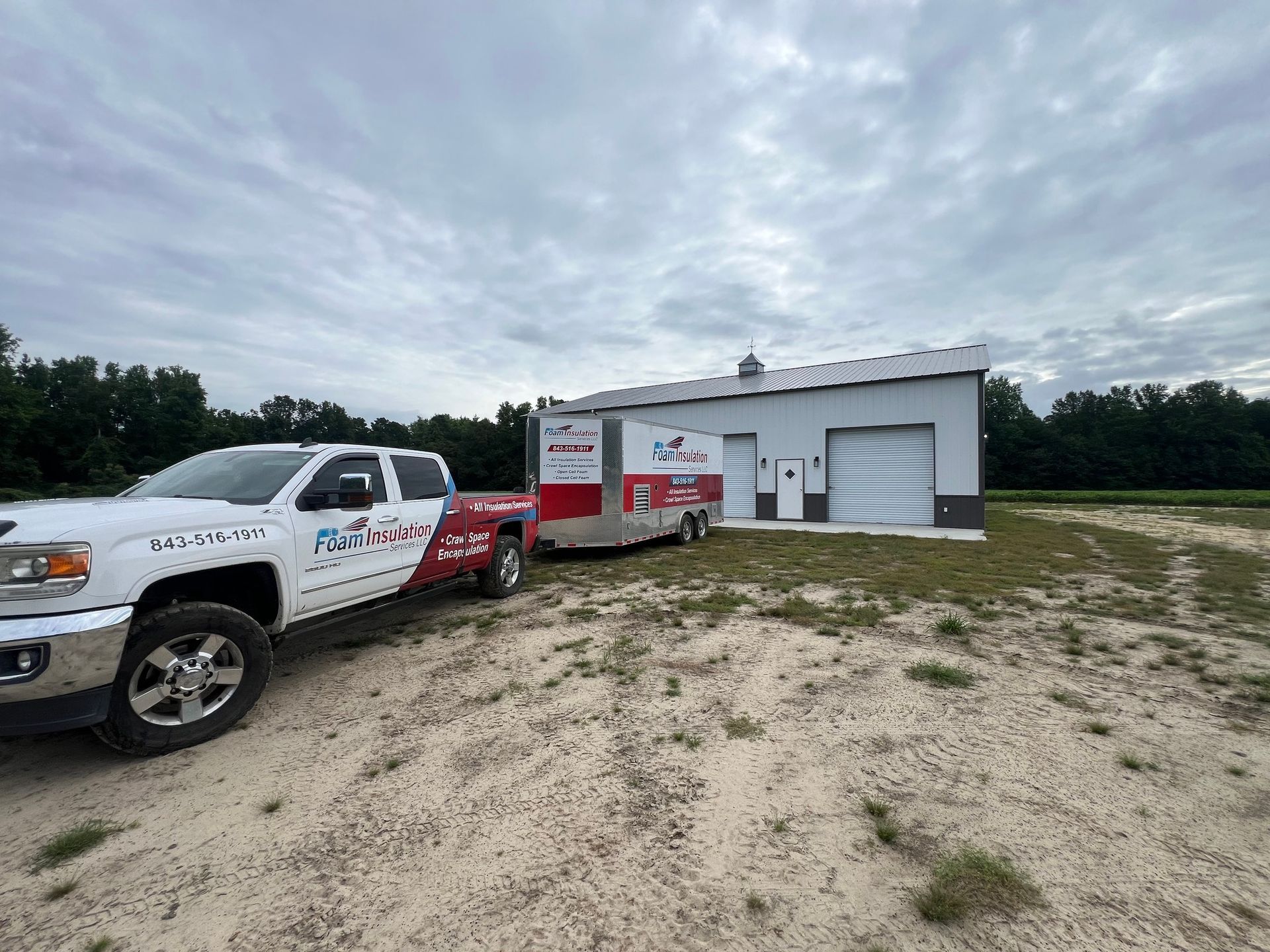 White truck towing a trailer parked near a white building with a garage door on a cloudy day.