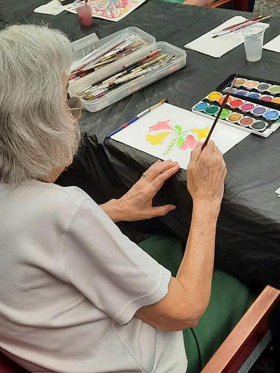 An elderly woman is sitting at a table painting with watercolors.