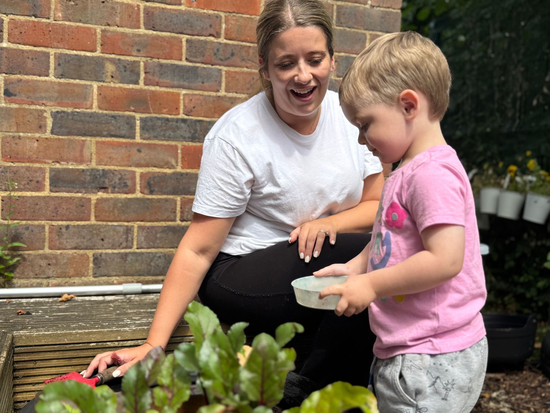 Toddler helping to water plants in nursery growing patch with Nursery Nurse