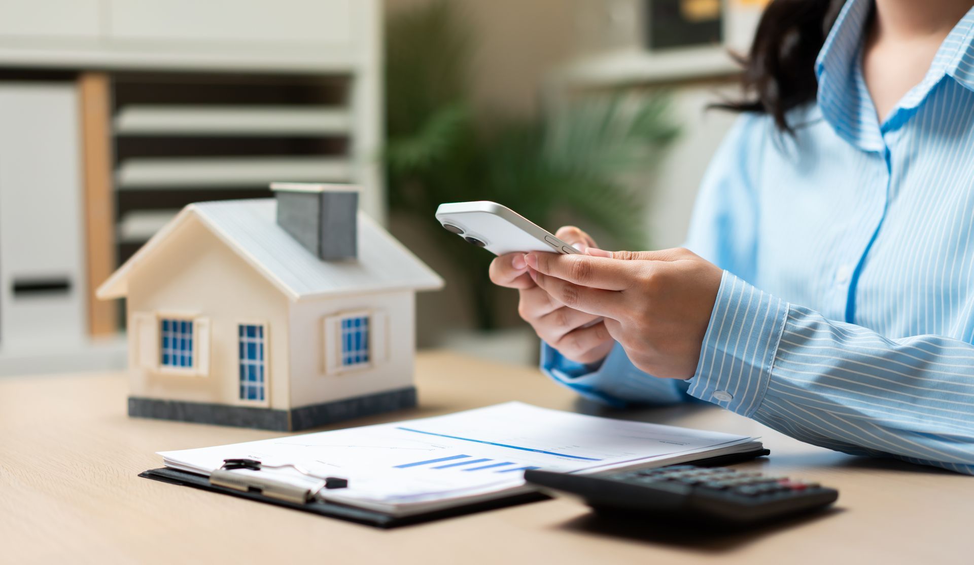 Person holding a smartphone over a desk with a small house model, clipboard with charts, and