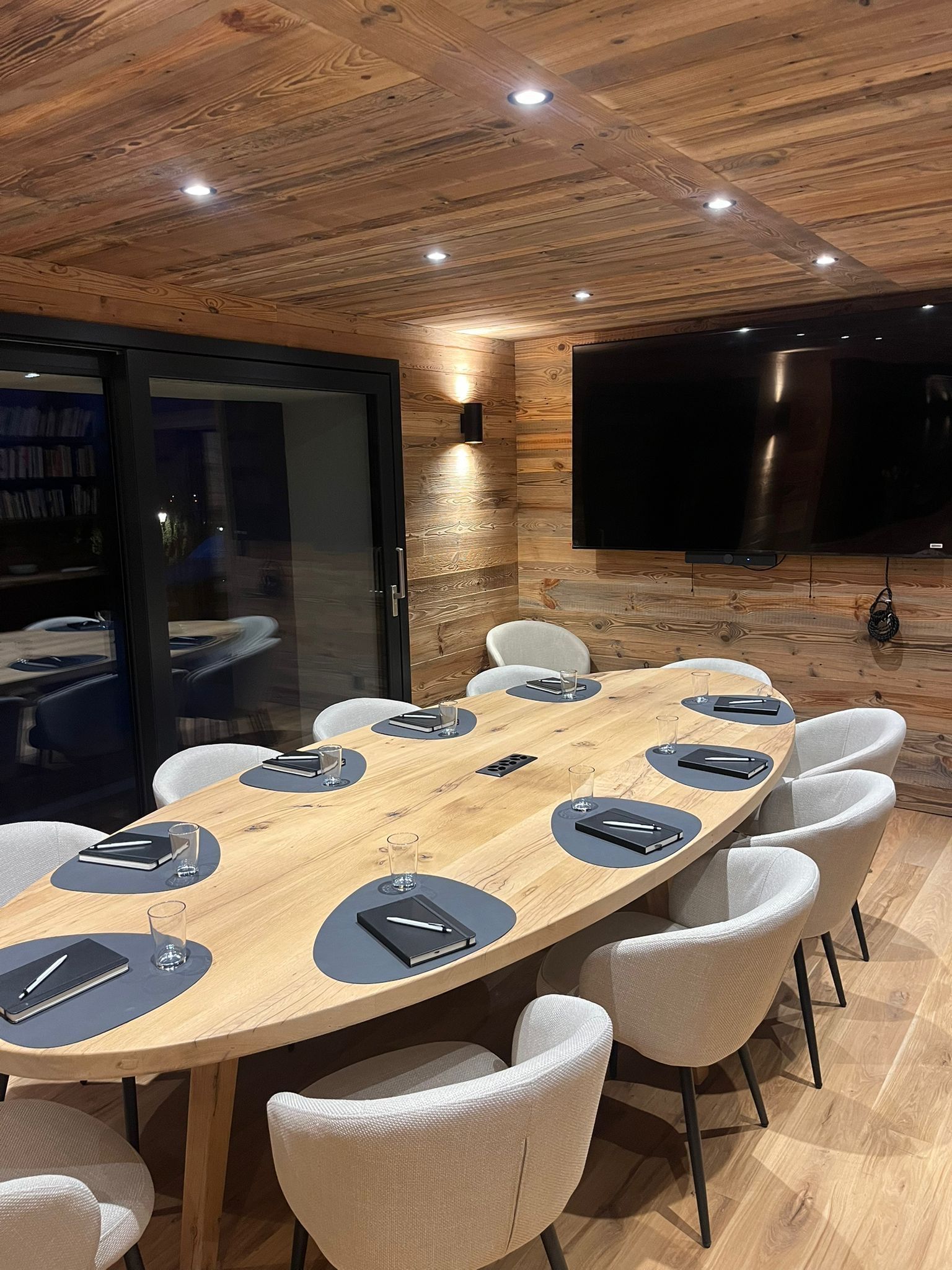 A wooden meeting table with chairs and notebooks in a room with wood-paneled walls and a ceiling with recessed lighting.