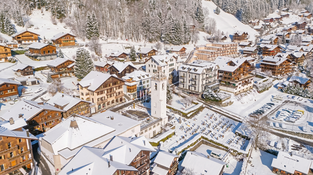 Vue aérienne d'un village suisse recouvert de neige, avec son clocher d'église imposant, ses maisons en bois et ses arbres denses.