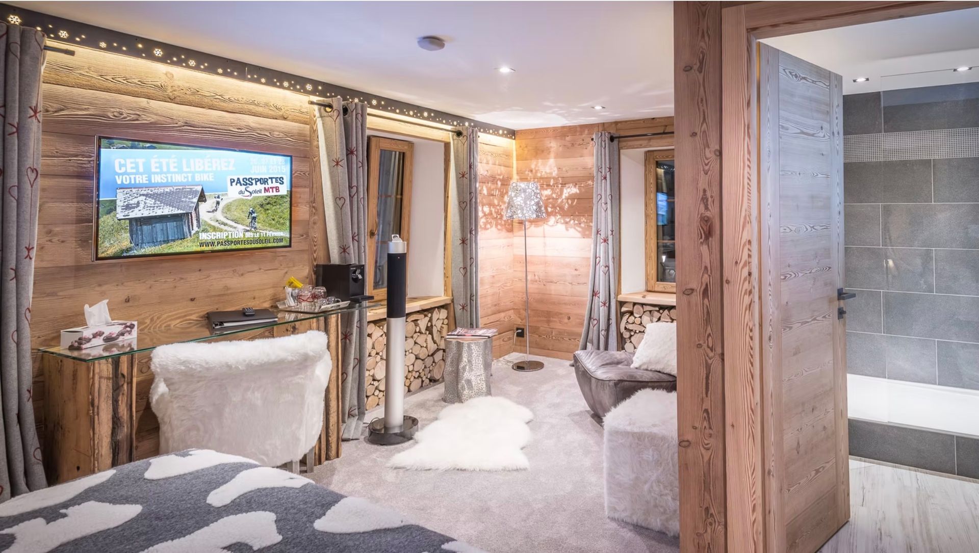 A cozy mountain-style bedroom with wood paneling, a wall-mounted TV, faux fur accents, and a view into a tiled bathroom.