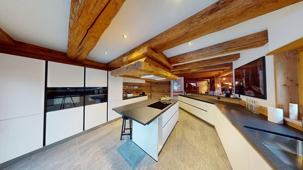 A modern, sleek kitchen with white cabinetry, a central island, and a dark stone countertop under exposed wooden beams.
